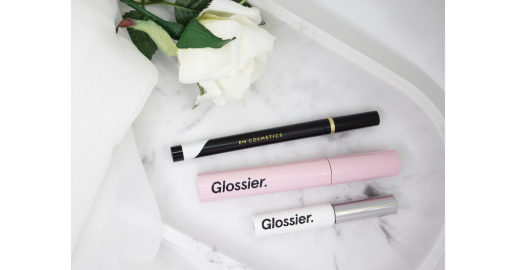 Makeup Products: Em Cosmetics Illustrated Eyeliner, Glossier Lash Slick Mascara, Glossier Boy Brow Brow Gel displayed in a white marble dish.