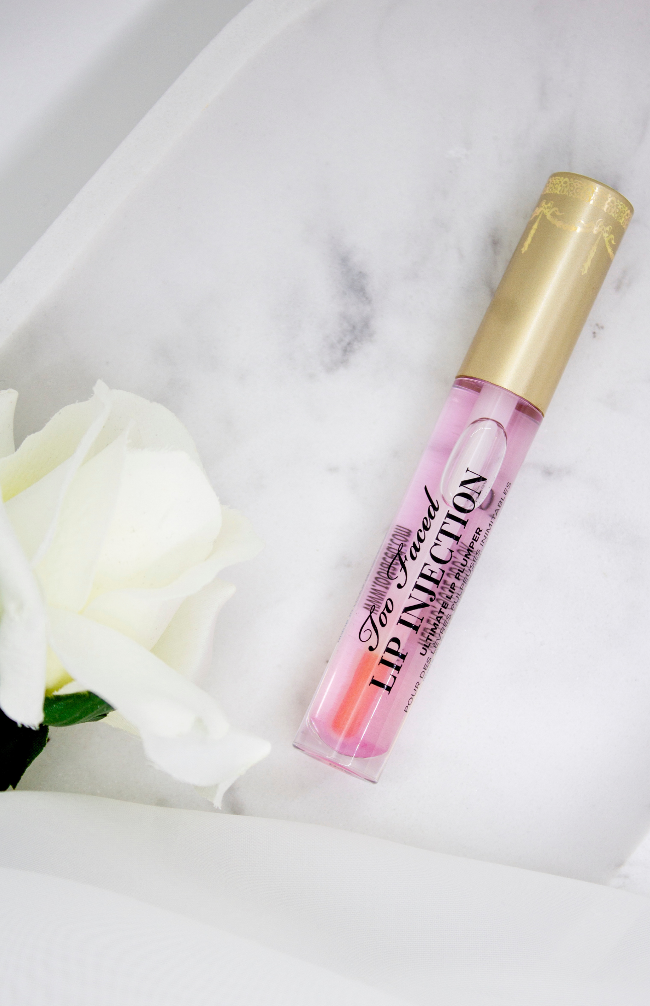 Makeup Products: Plump and Pout with Too Faced Lip Plumper - Presented on a White Marble Dish, Adorned with a Delicate White Rose. Elevate Your Beauty Game!