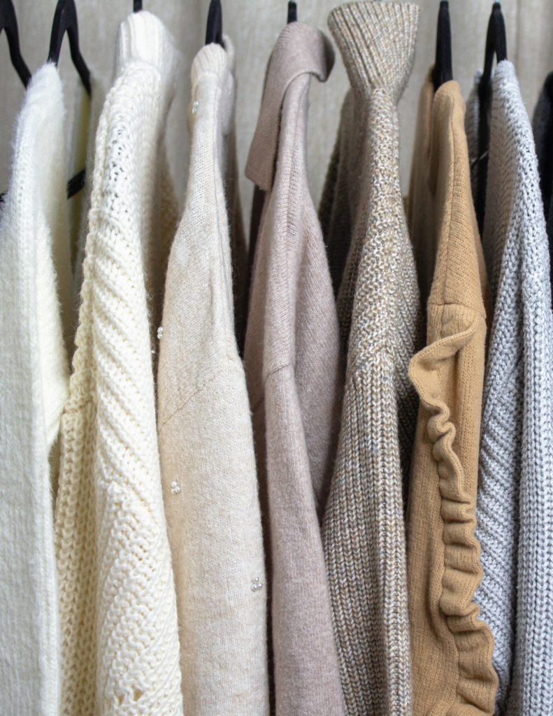 In the image, a clothing rack displays an array of luxurious cashmere and sophisticated fine-knit sweaters. The collection includes various colors such as white, cream, beige, light brown, mustard, and light blue, embodying quiet luxury, timeless elegance, and classic, neutral color palette.