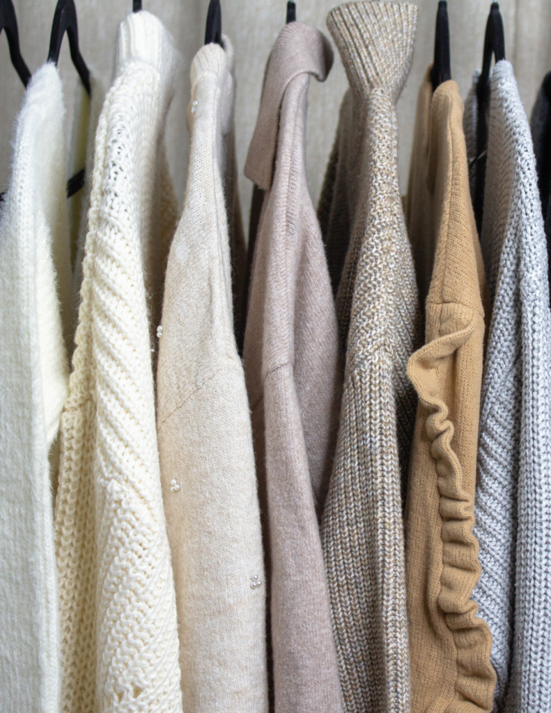 Fall wardrobe essentials on a clothing rack featuring luxurious cashmere and sophisticated fine-knit sweaters in colors like white, cream, beige, light brown, mustard, and light blue, embodying quiet luxury and timeless elegance.