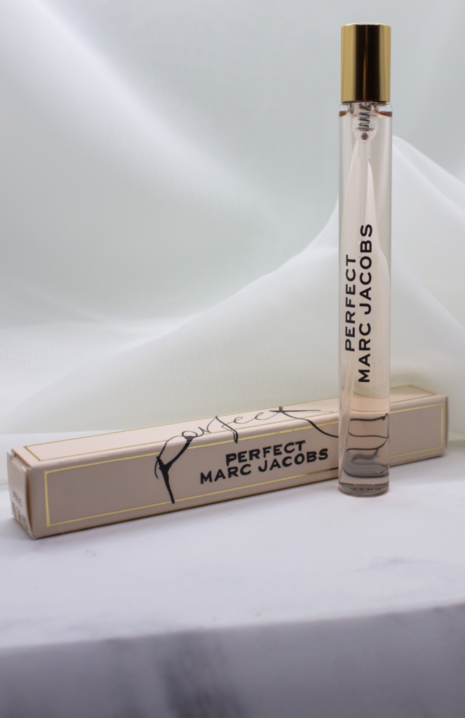 Travel-size Marc Jacobs fragrance bottle with its packaging displayed on a marble platform against a soft neutral background. The simple setup showcases the fragrance in a clean and stylish presentation.